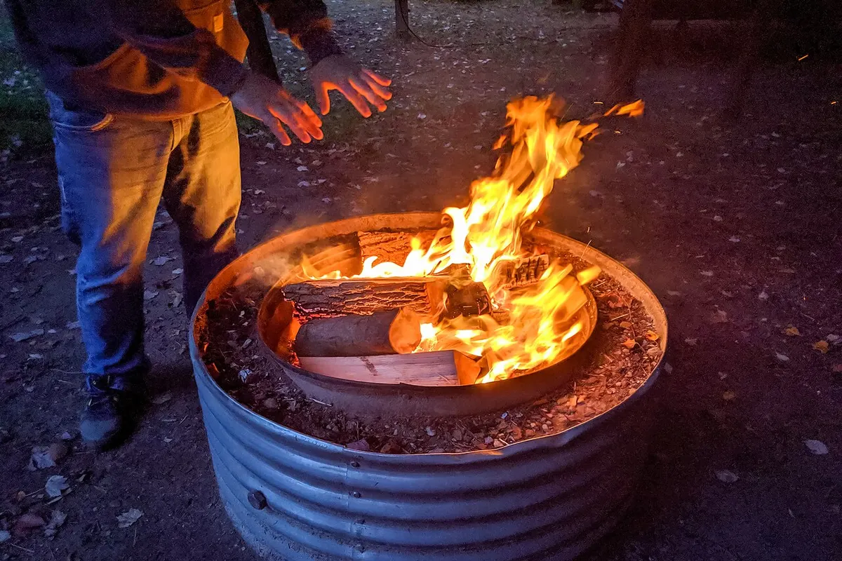 A campfire burning in a metal ring with a person warming their hands nearby