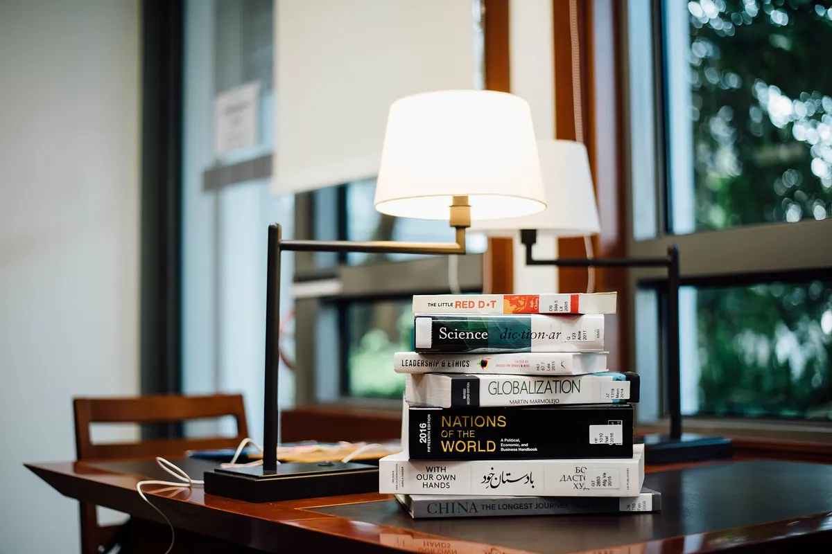 A stack of books and a reading lamp on a desk by a window